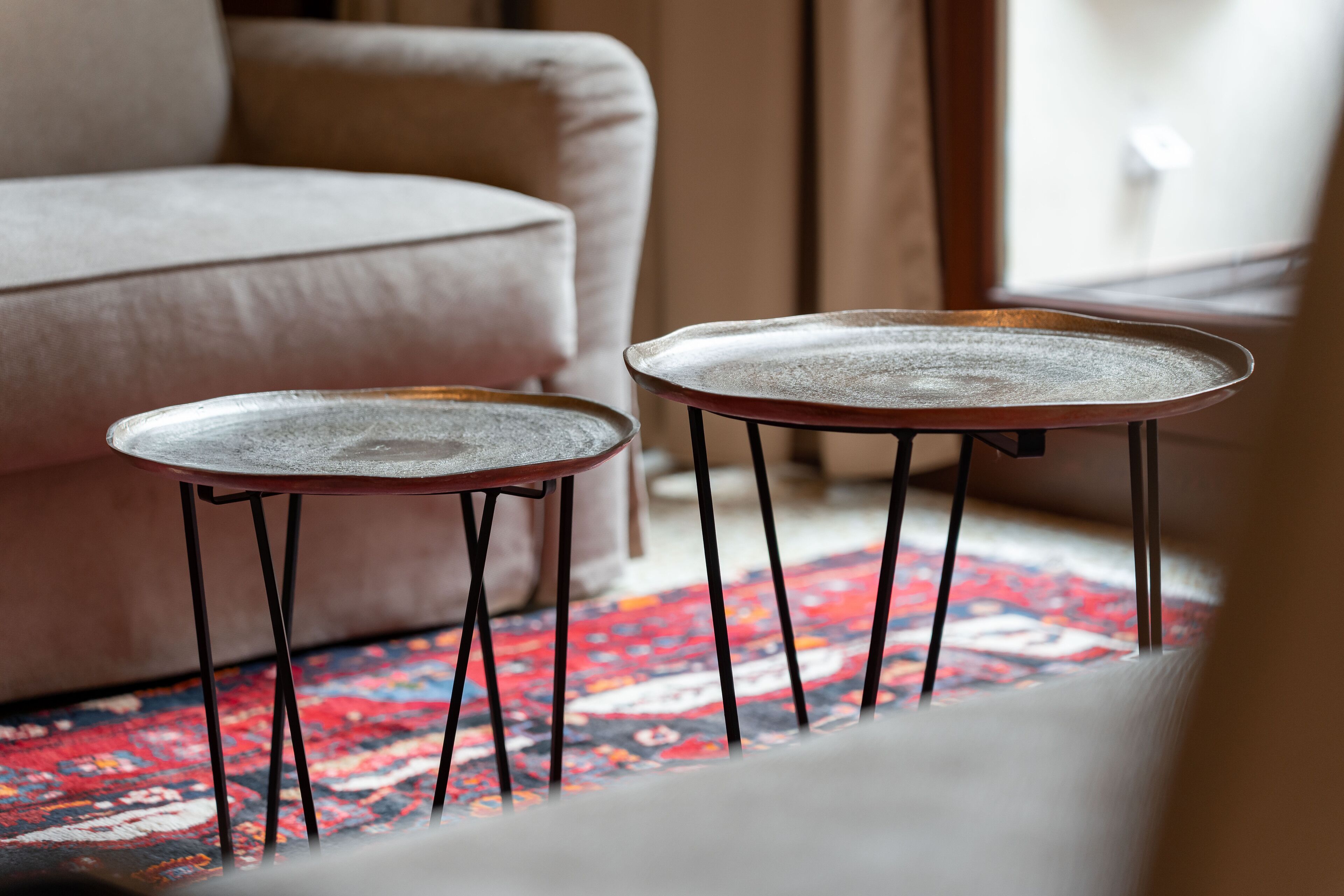 Deux petites tables rondes aux pieds en métal, posées sur un tapis coloré dans une pièce où se trouve un canapé beige.