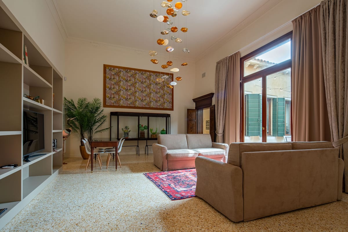 A living room with a sofa, a coffee table, a dining table with chairs, a bookshelf, a large potted plant, and a colorful rug.