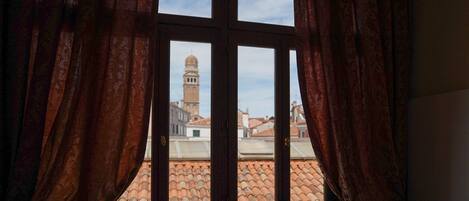 A window with a view of a building with a dome and a bell tower, curtains, and a tiled roof.