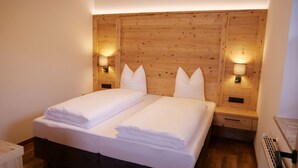 Cama doble con sábanas blancas, cabecera de madera y dos mesitas de noche fijadas a la pared con lámparas.