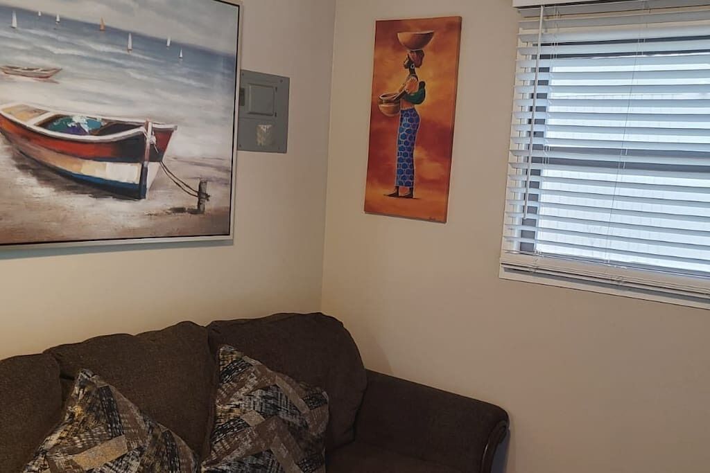 Ein Wohnzimmer mit braunem Sofa, einem Gemälde von einem Boot auf dem Wasser und einem weiteren Gemälde, das eine Person mit einem Topf zeigt.