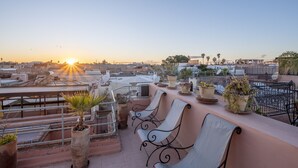 Een terras op het dak met zitplaatsen, plantenpotten en een uitzicht op de ondergaande zon boven een stadsbeeld.