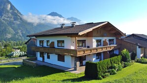 Een chalet met twee verdiepingen en een balkon, omgeven door weelderige begroeiing en bergen op de achtergrond.