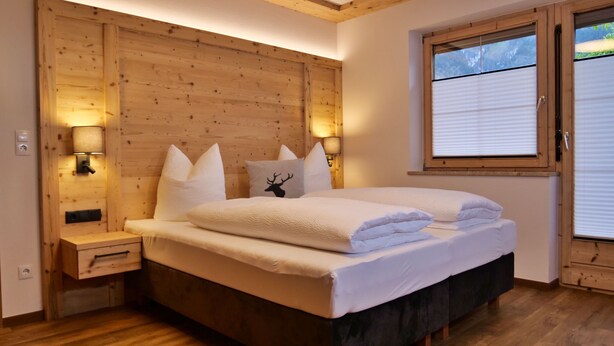 Een slaapkamer met een houten hoofdeinde, een bed met witte lakens, twee nachtlampjes en een raam met jaloezieën.