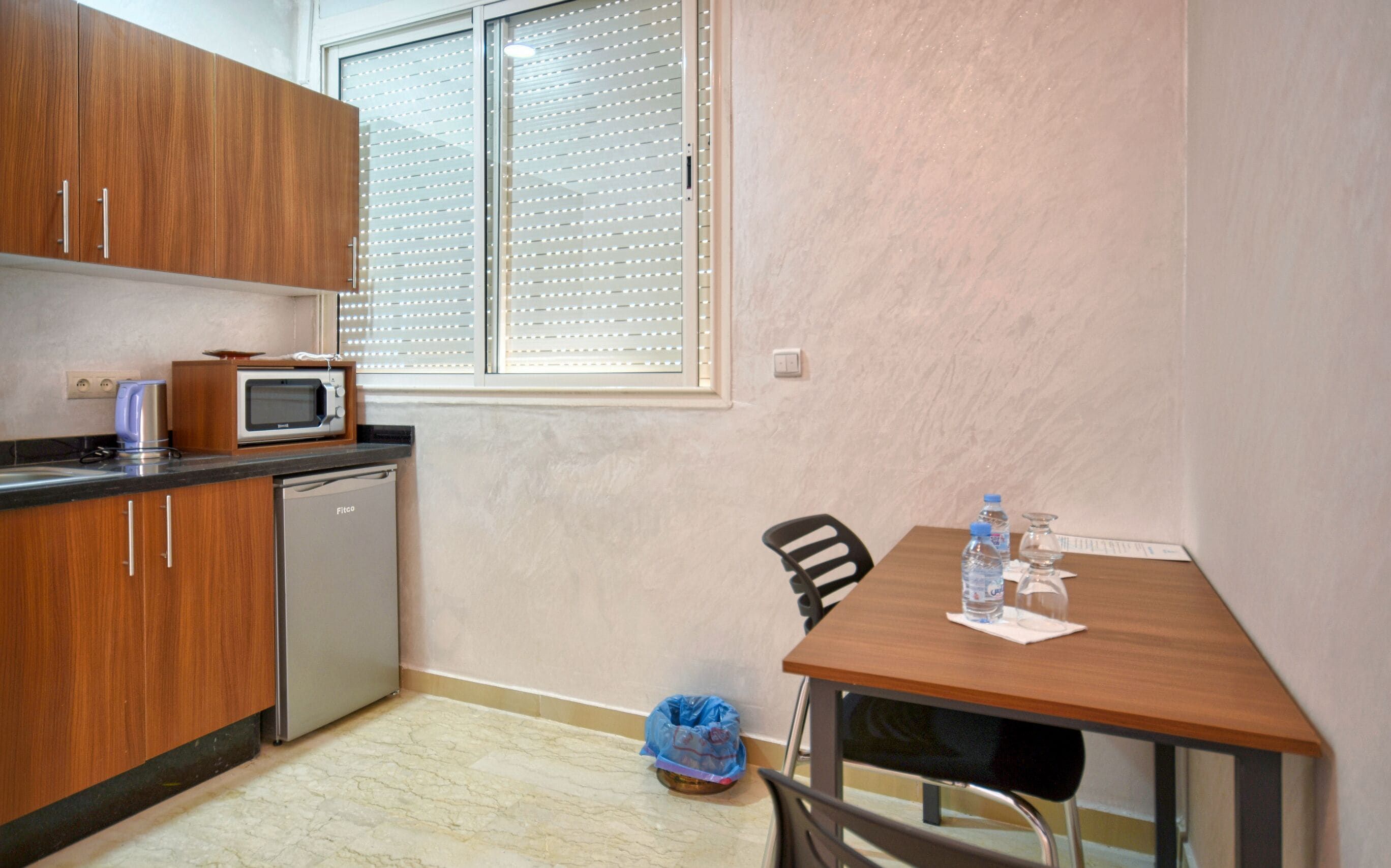 A compact kitchen with wooden cabinets, a microwave, and a small refrigerator. There's a dining table with a chair and a water bottle on it.