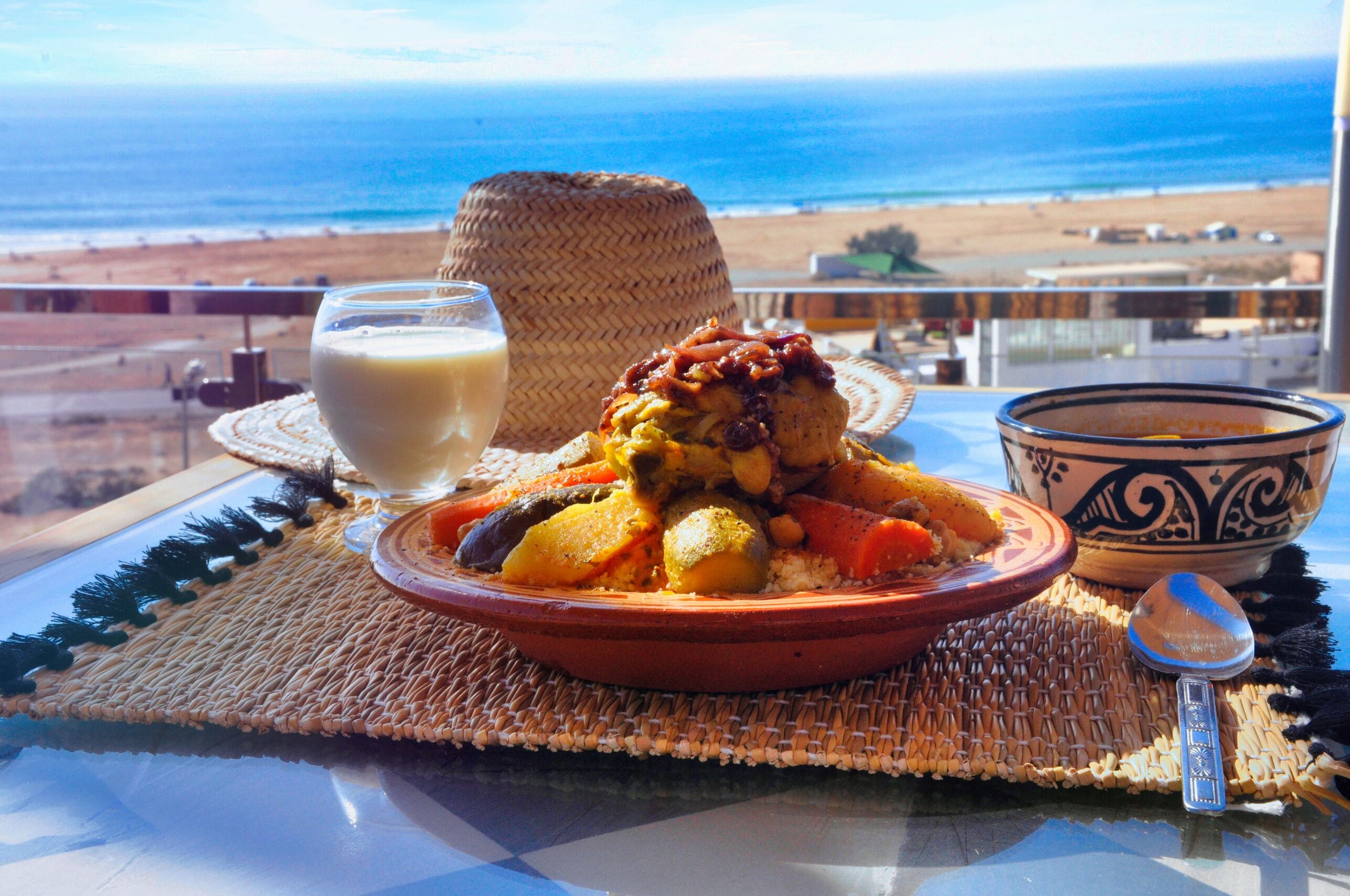 A meal of couscous with vegetables and meat, a glass of milk, and a woven basket on a table with a view of the beach and ocean.