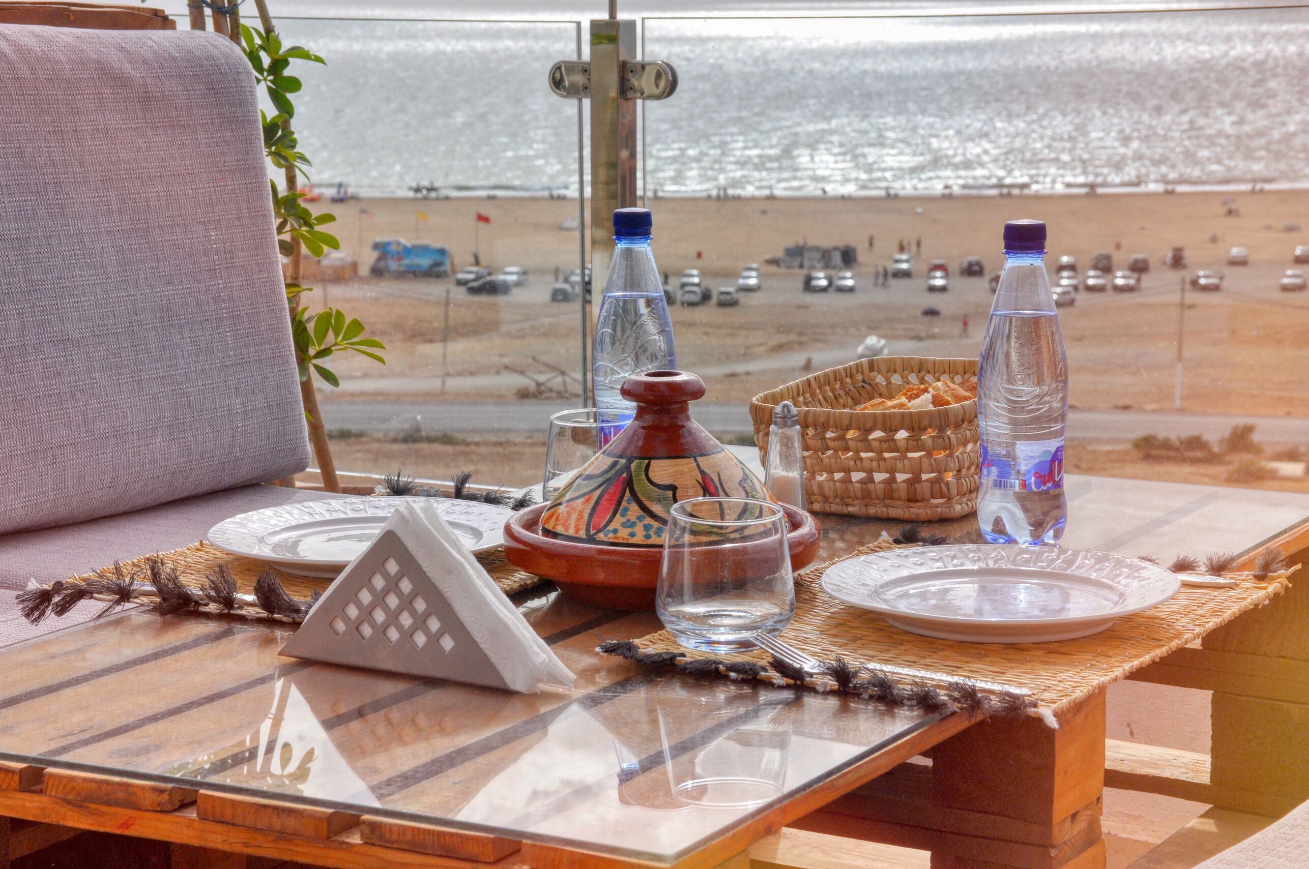 A table set with plates, glasses, and a water bottle, overlooking a beach with parked cars and people.