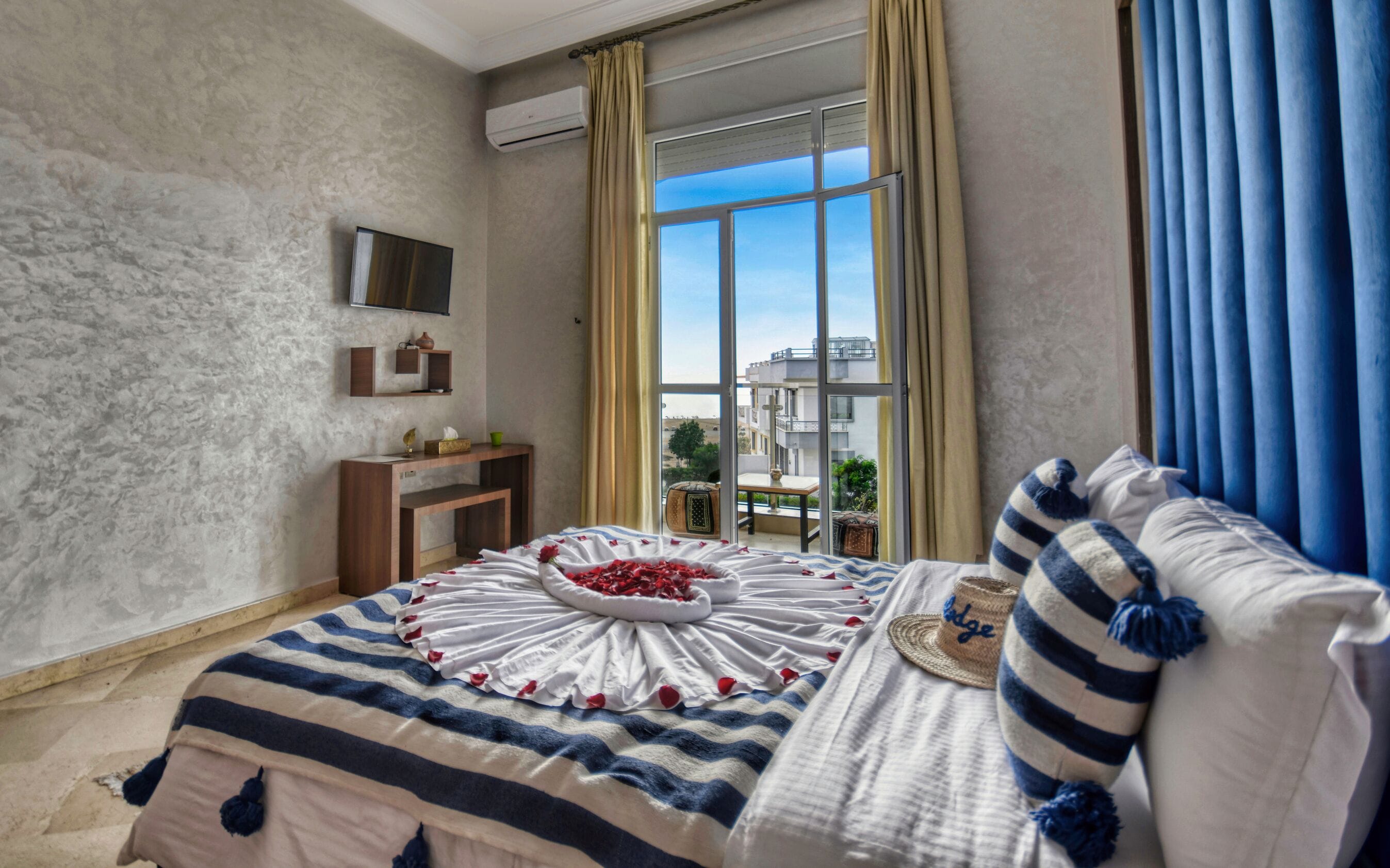 A bedroom with a large bed, a bedside table, a mounted TV, and a view of buildings through a window.