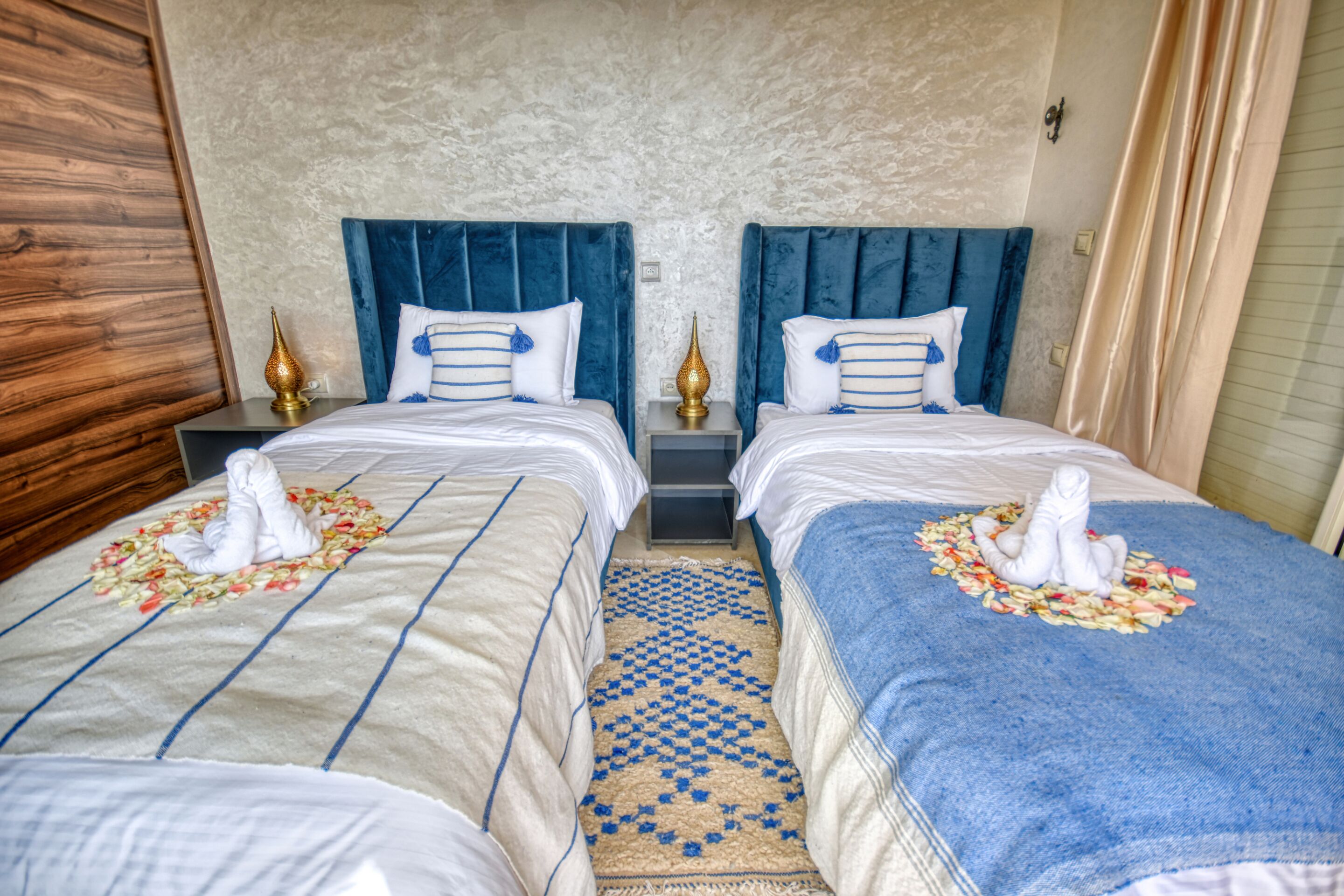 A hotel room with two beds, each with a blue headboard, white bedding, and a decorative towel arrangement.