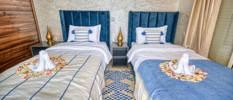 A hotel room with two beds, each with a blue headboard, white bedding, and a decorative towel arrangement.