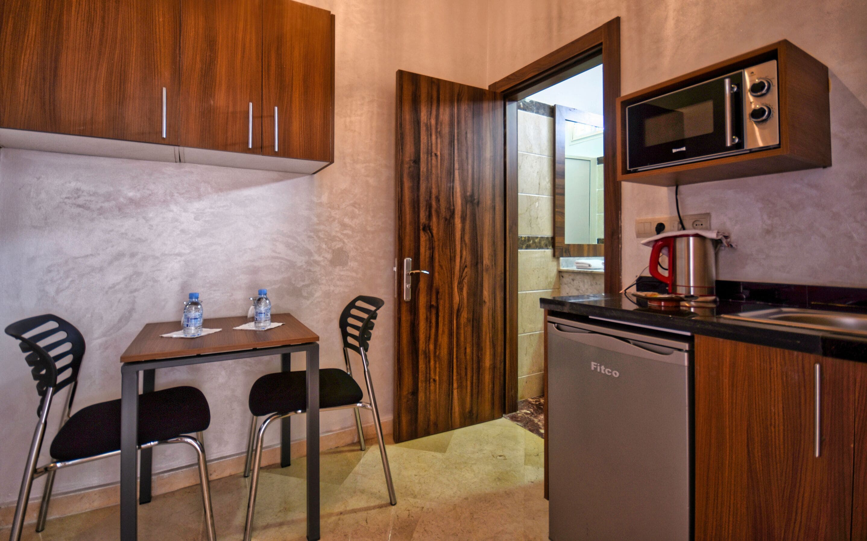 A compact kitchen with wooden cabinets, a microwave, a coffee maker, and a dishwasher. There is a small table with two chairs and two water bottles on it.