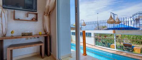 Habitación con televisión, un banco de madera, una pequeña estantería con objetos decorativos y vistas a una piscina y zona exterior.