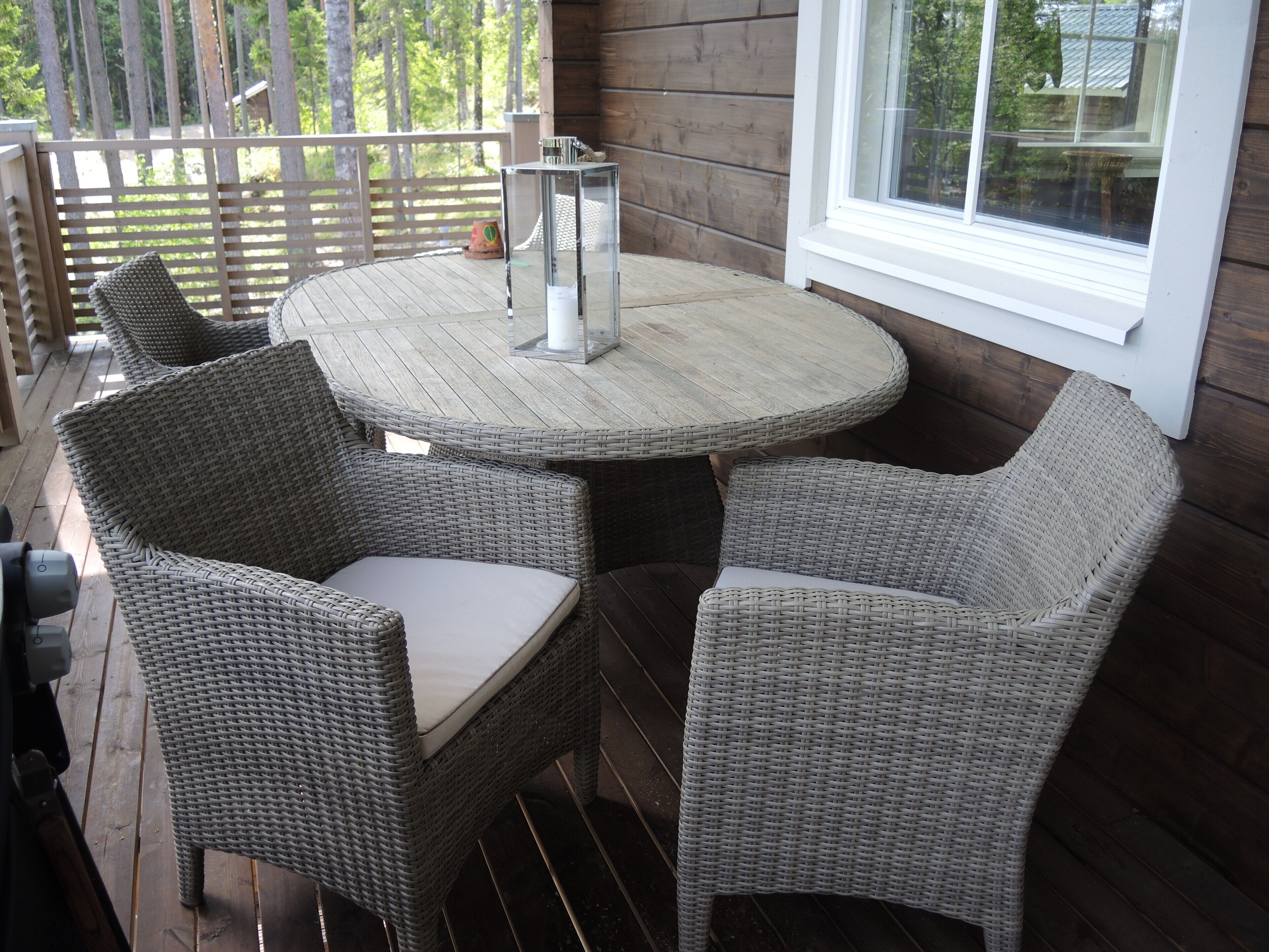 A wicker table and chairs on a wooden deck with a window and railing.