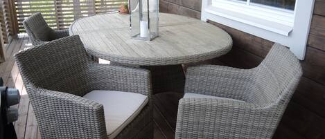 A wicker table and chairs on a wooden deck with a window and railing.