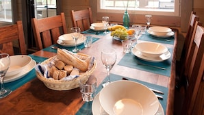 A wooden dining table set with plates, glasses, a fruit bowl, and bread.