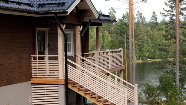 A wooden cabin with a wooden staircase leading to a balcony, overlooking a lake surrounded by trees.