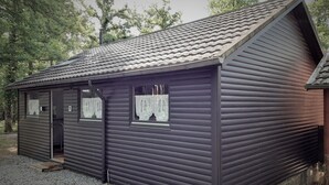 A small, dark wooden cabin with a tiled roof, a door, and windows with lace curtains.