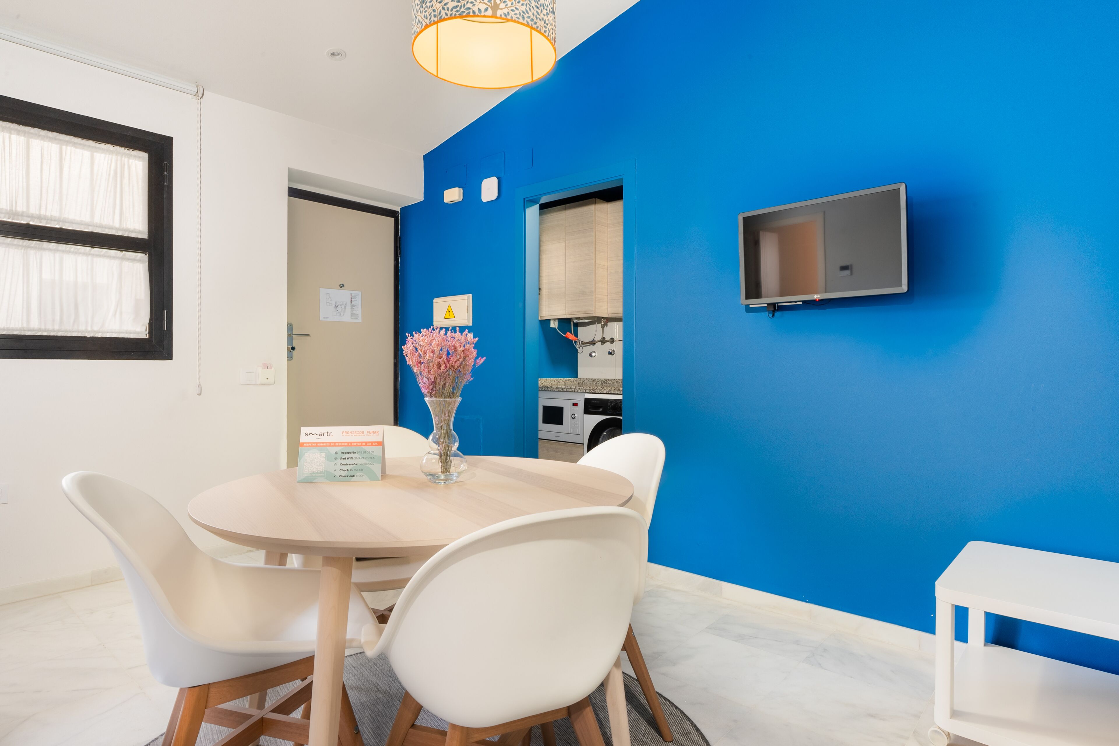Ein modernes Zimmer mit blauer Akzentwand, einem runden Tisch mit Stühlen, einer Vase mit Blumen, einem an der Wand montierten Fernseher und einem kleinen weißen Regal.