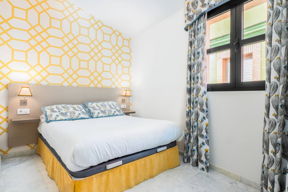 A neatly made bed with a white duvet and patterned pillows, a geometric wall design, and a window with curtains.