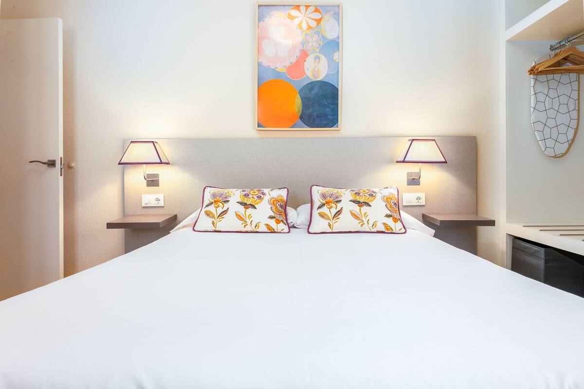 A neatly made bed with floral pillows, two bedside lamps, and a wall art piece above.