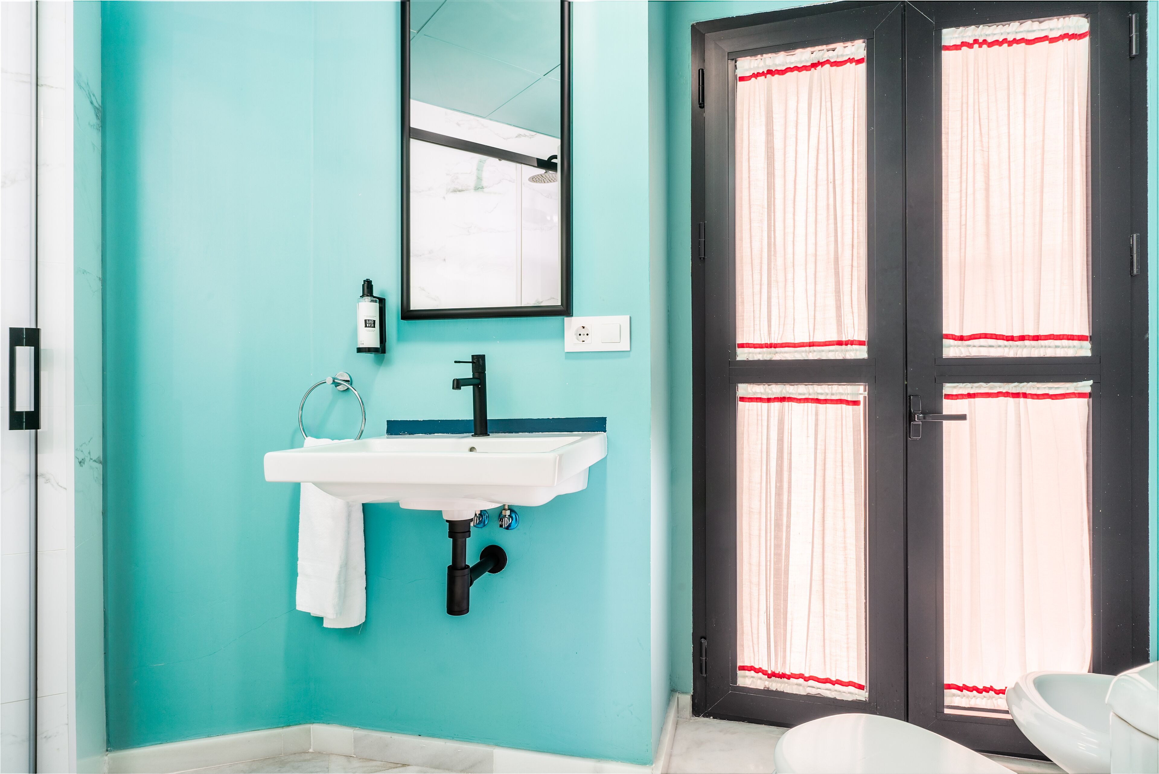 Un bany amb una paret color turquesa, un lavabo blanc, un mirall i una porta amb cortines vermelles.