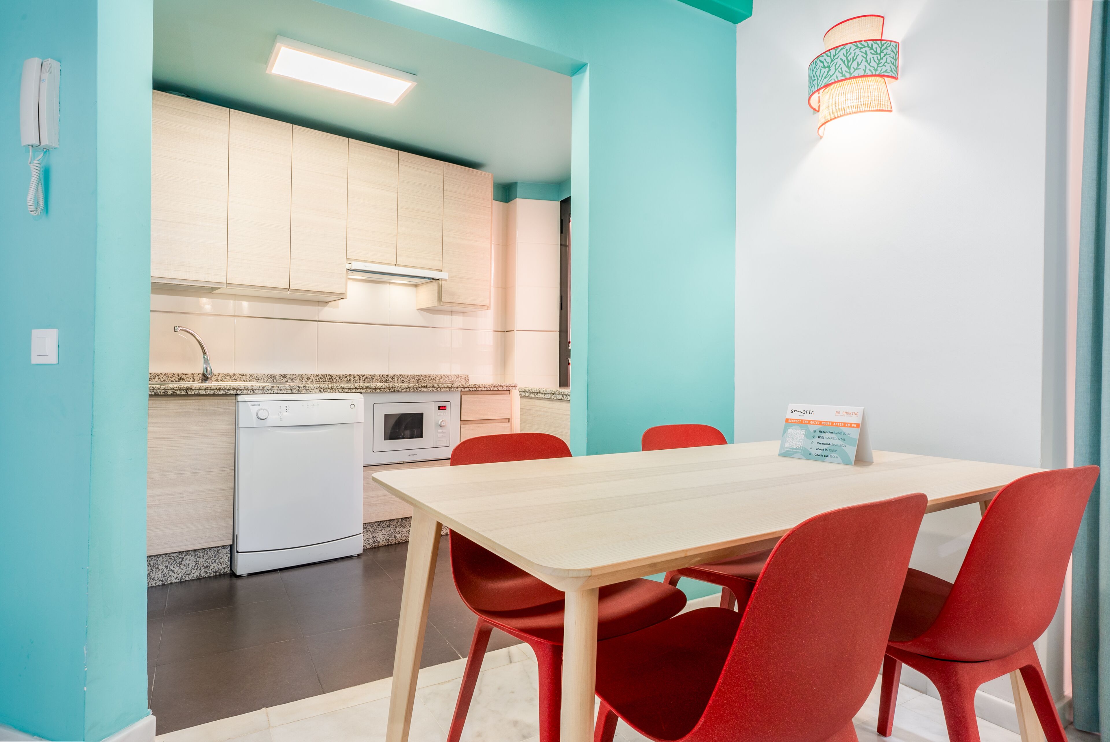 A compact kitchen with a dishwasher, microwave, and a small oven. There is a table with four red chairs and a light fixture on the wall.