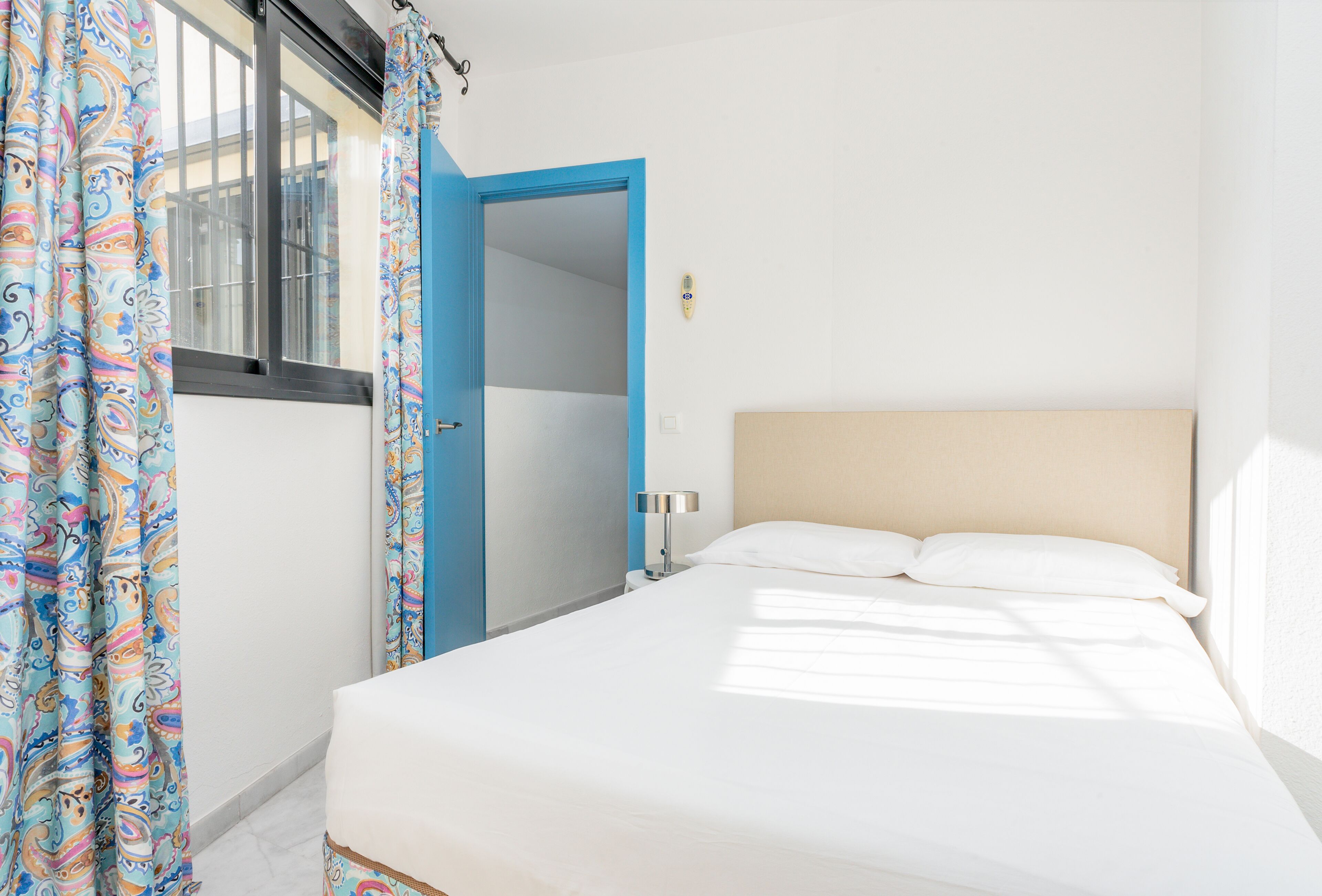 A bedroom with a bed, a nightstand, a blue door, and a window with curtains.