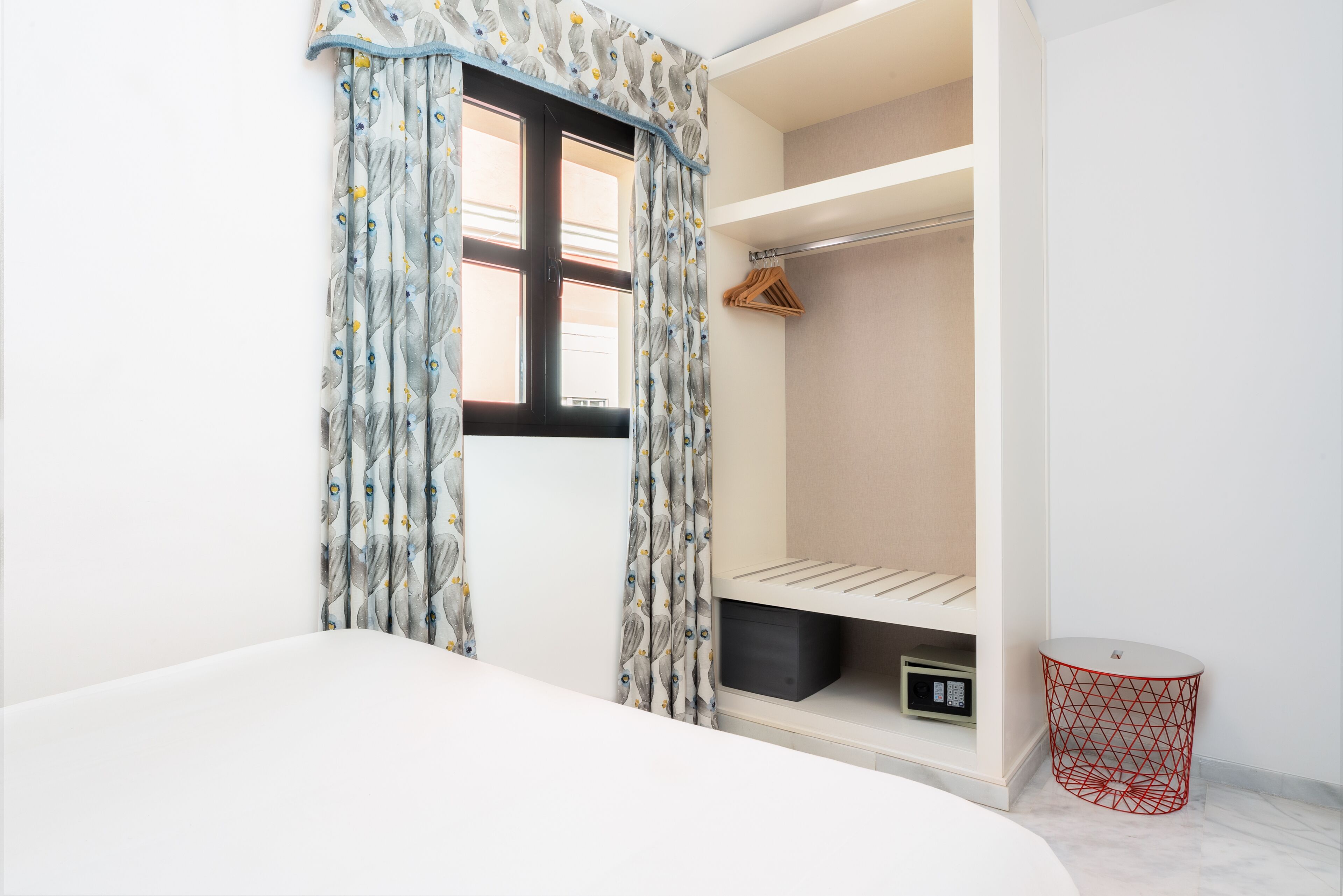 A bedroom with a built-in closet, a window with patterned curtains, and a small red wire basket.