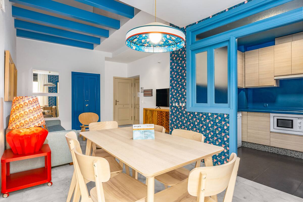 A modern kitchen with blue accents, a dining area with a wooden table and chairs, and a red chair with a patterned cushion.