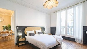 A hotel room with a large bed, a bedside table with a lamp, a chair, and a view of the city through large windows. - Le Champagne Central (Reims)