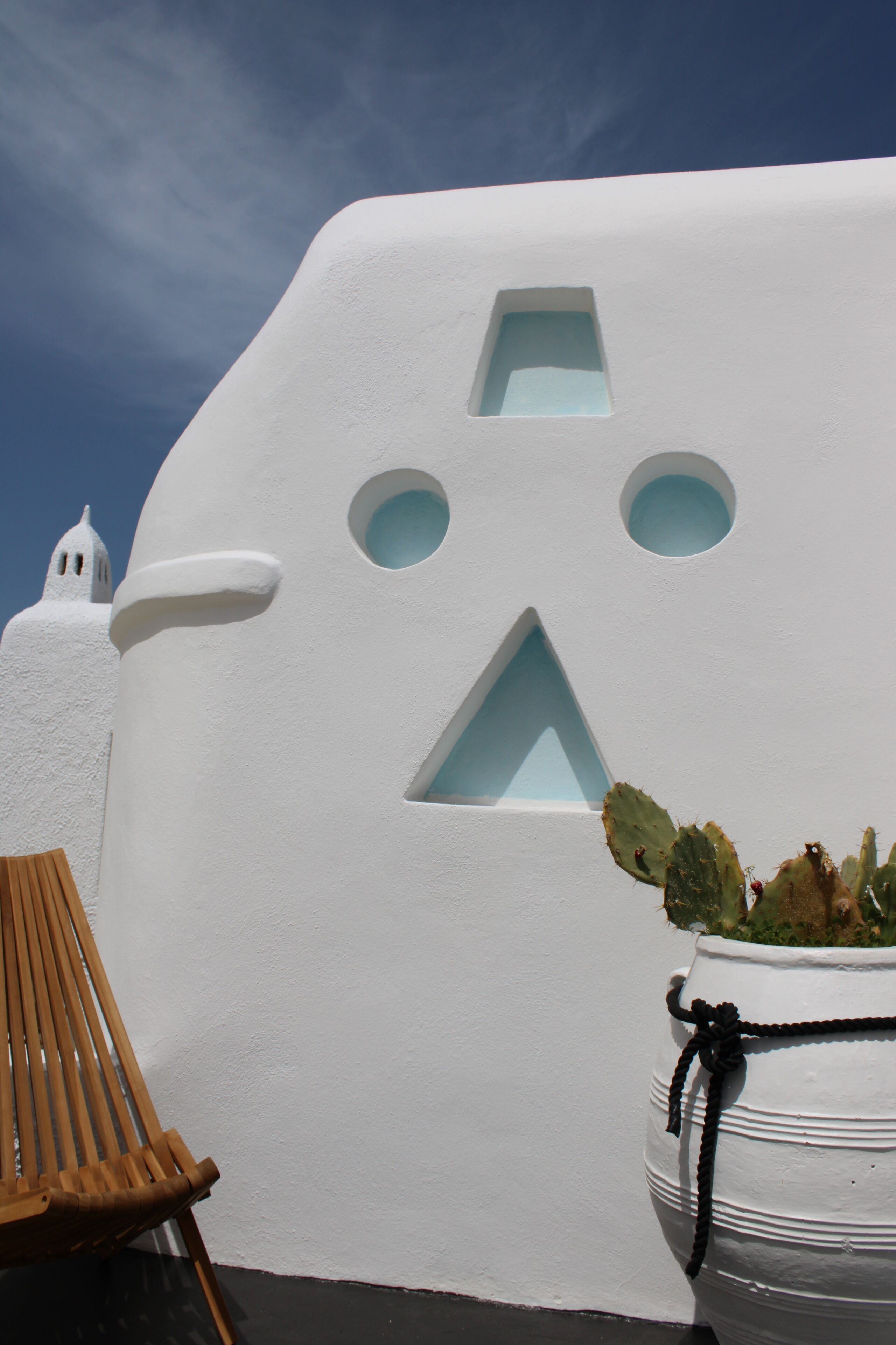 A white building with a unique geometric facade, a wooden chair, and a potted plant.