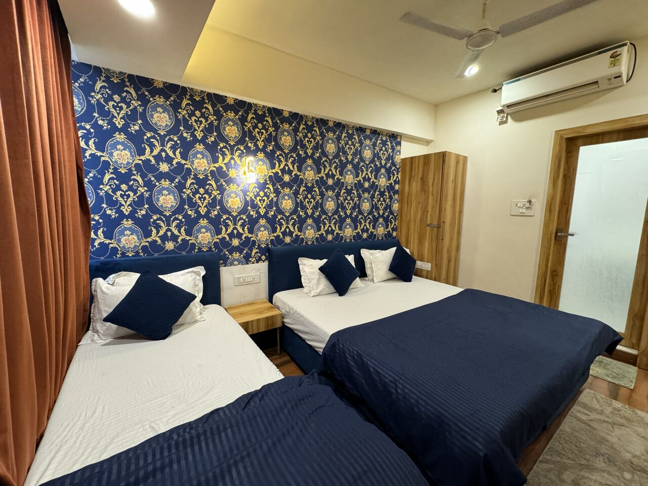 A hotel room with two beds, a wooden wardrobe, a nightstand, and a patterned wallpaper.