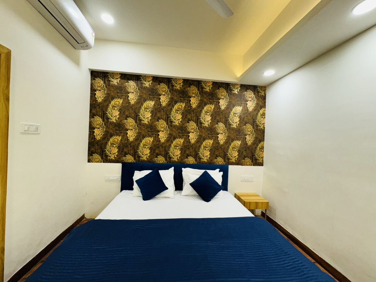 A neatly made bed with a dark blue bedspread and white pillows, a wooden headboard with a floral pattern, and a small bedside table with a lamp.