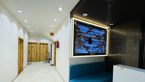 A hallway with a blue bench, wooden doors, and a wall-mounted screen displaying a scenic image. - Hotel Shree Mahakal Krapa (Ujjain)