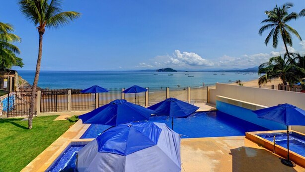 A pool area with blue umbrellas, a clear view of the ocean, and palm trees. - Casa las Mariposas Bungalow 9 (Los Ayala)