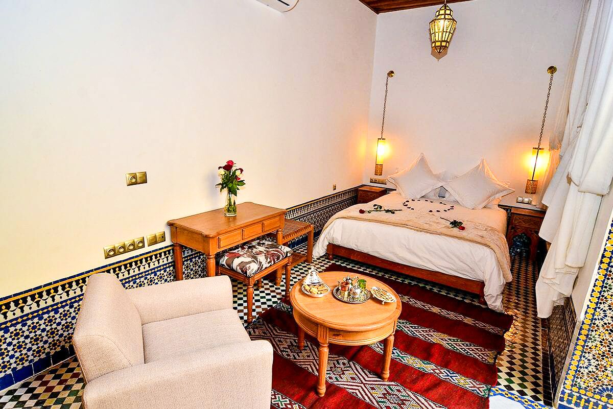 Quarto tradicional marroquino com uma cama, um sofá, uma pequena mesa com um vaso e ladrilhos no chão com padrões.