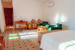 Quarto de hotel com uma cama, uma escrivaninha, um sofá e uma cadeira. Há uma janela com luz solar entrando.
