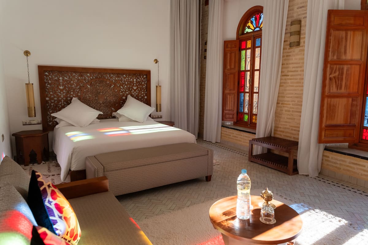 Ein Hotelzimmer mit einem großen Bett, einem hölzernen Kopfteil, einem kleinen Tisch mit Flasche und Glas sowie Blick auf ein Buntglasfenster.