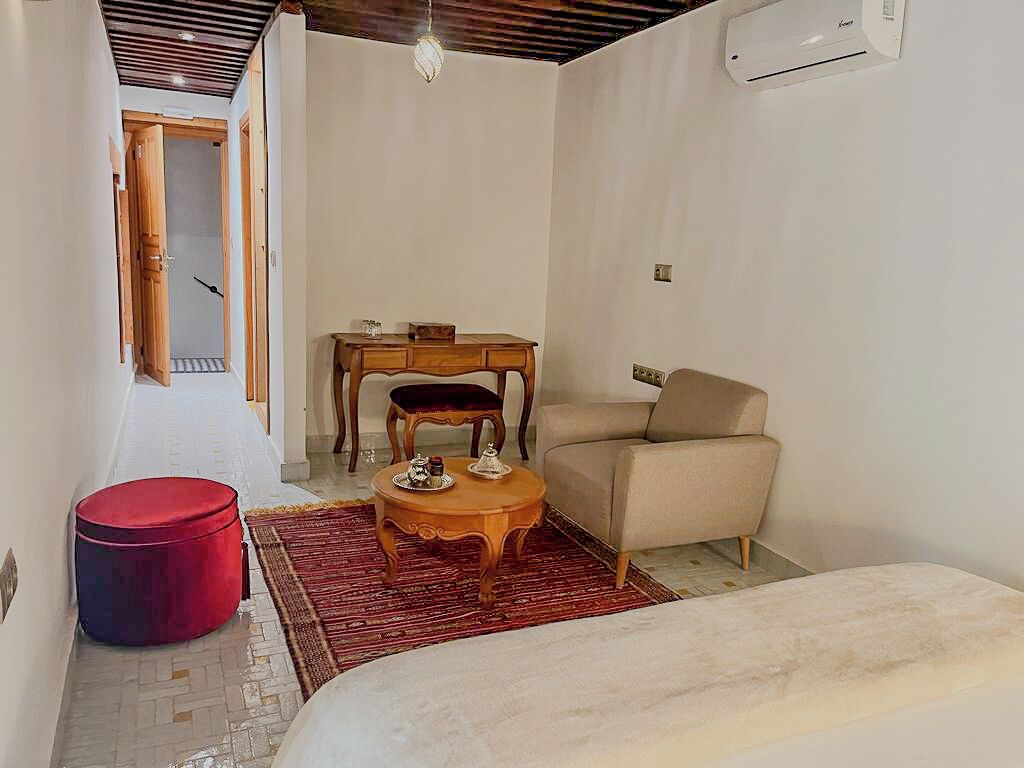 A compact room with a bed, a small round table with chairs, a sofa, and a red ottoman. There is a wooden desk with a chair and a small table with a lamp.