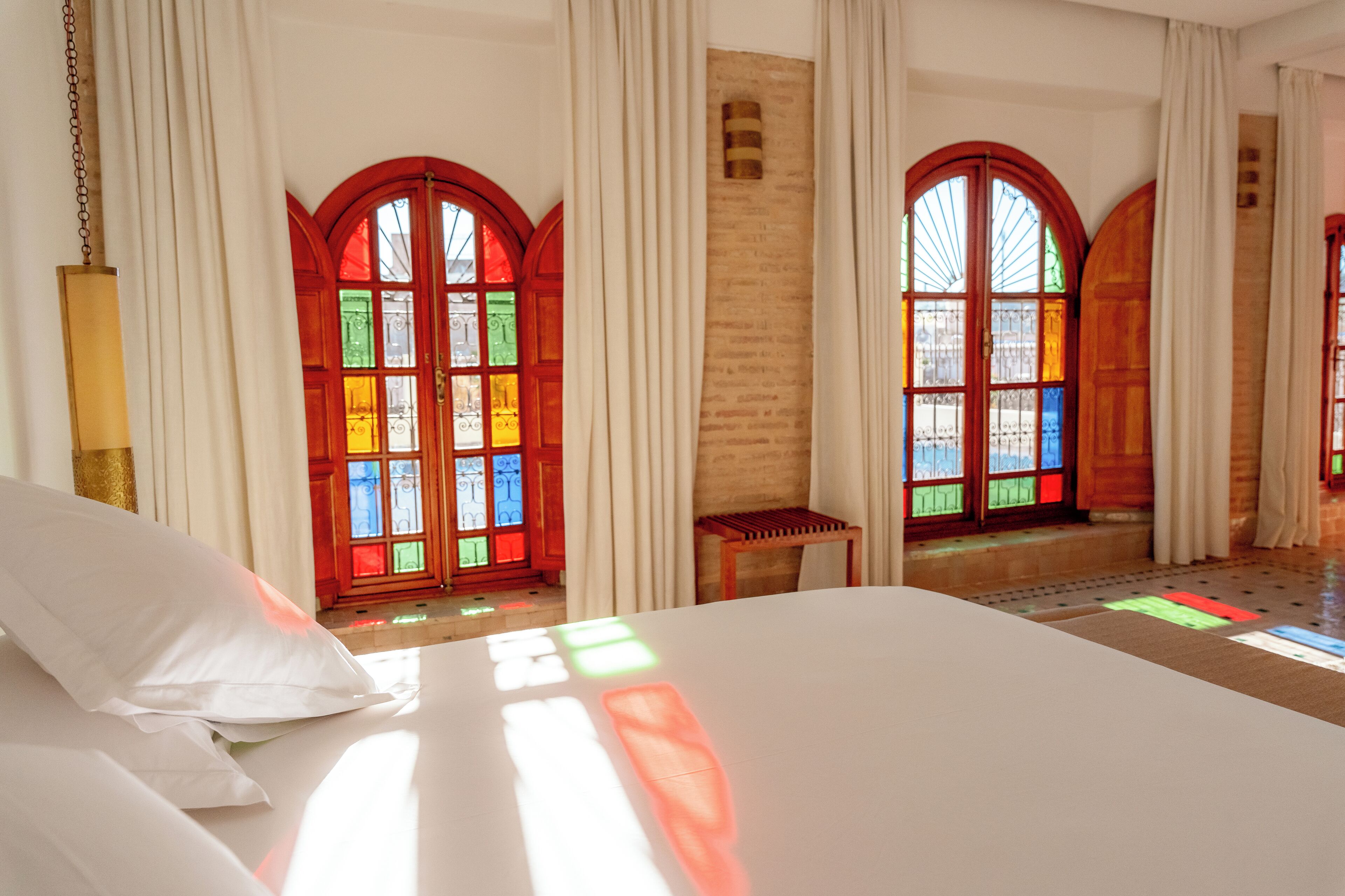 A hotel room with a large bed, two windows with stained glass, and a wooden bench.