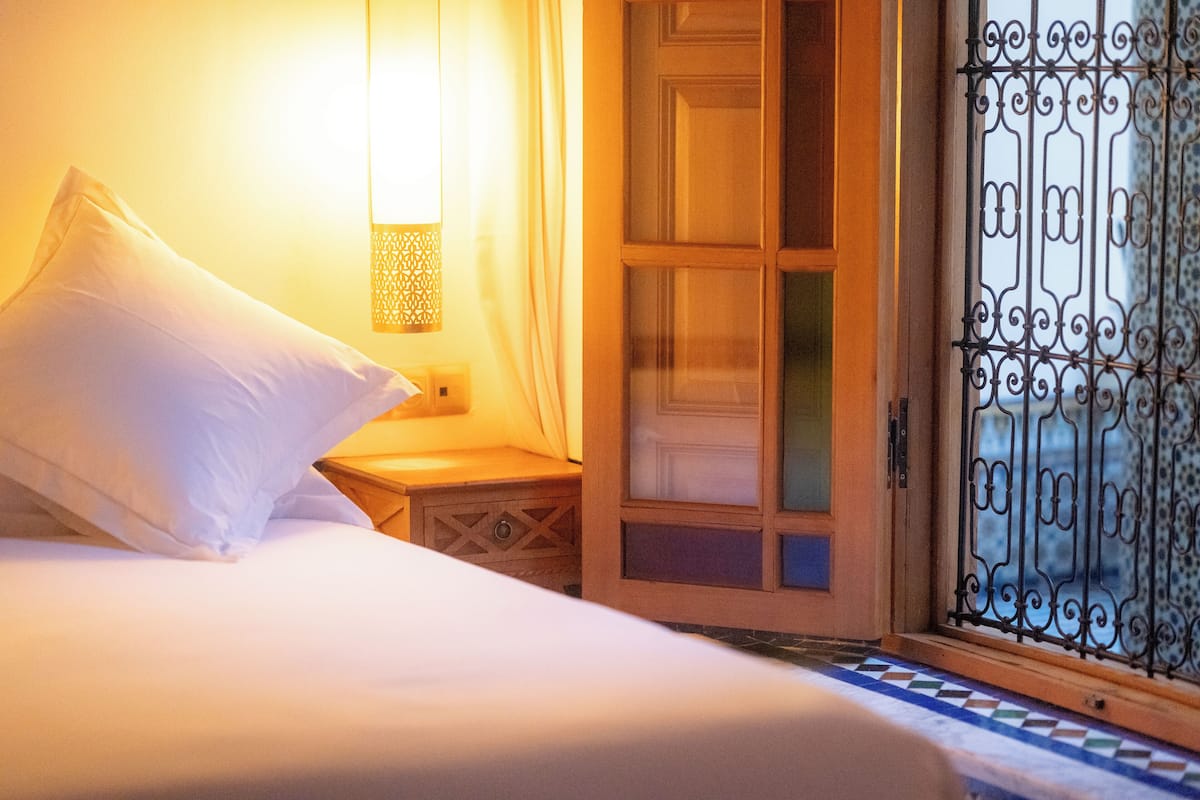 Ein Bett mit weißer Bettwäsche, ein Nachttisch, eine Wandlampe und ein Fenster mit schmiedeeisernen Gitterstäben.
