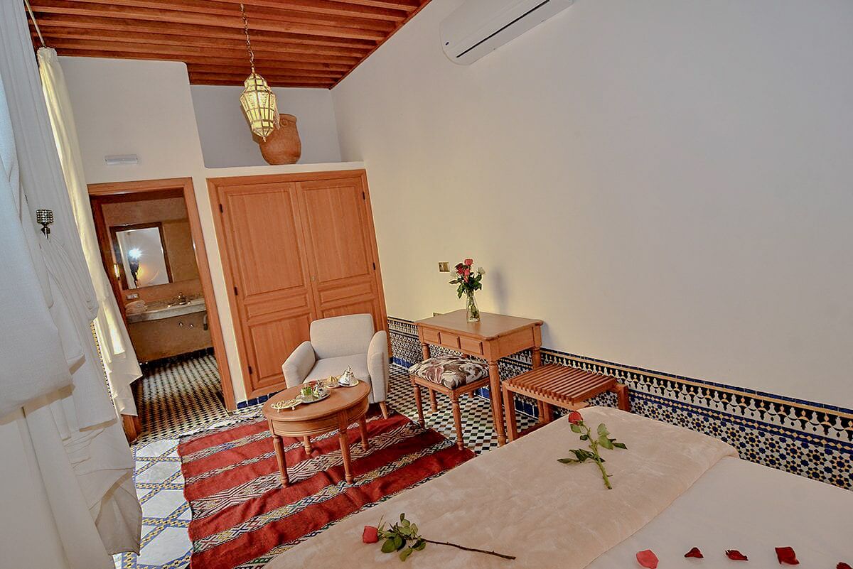 Una habitación con una cama, una silla de madera, una mesita con un florero y una alfombra estampada.