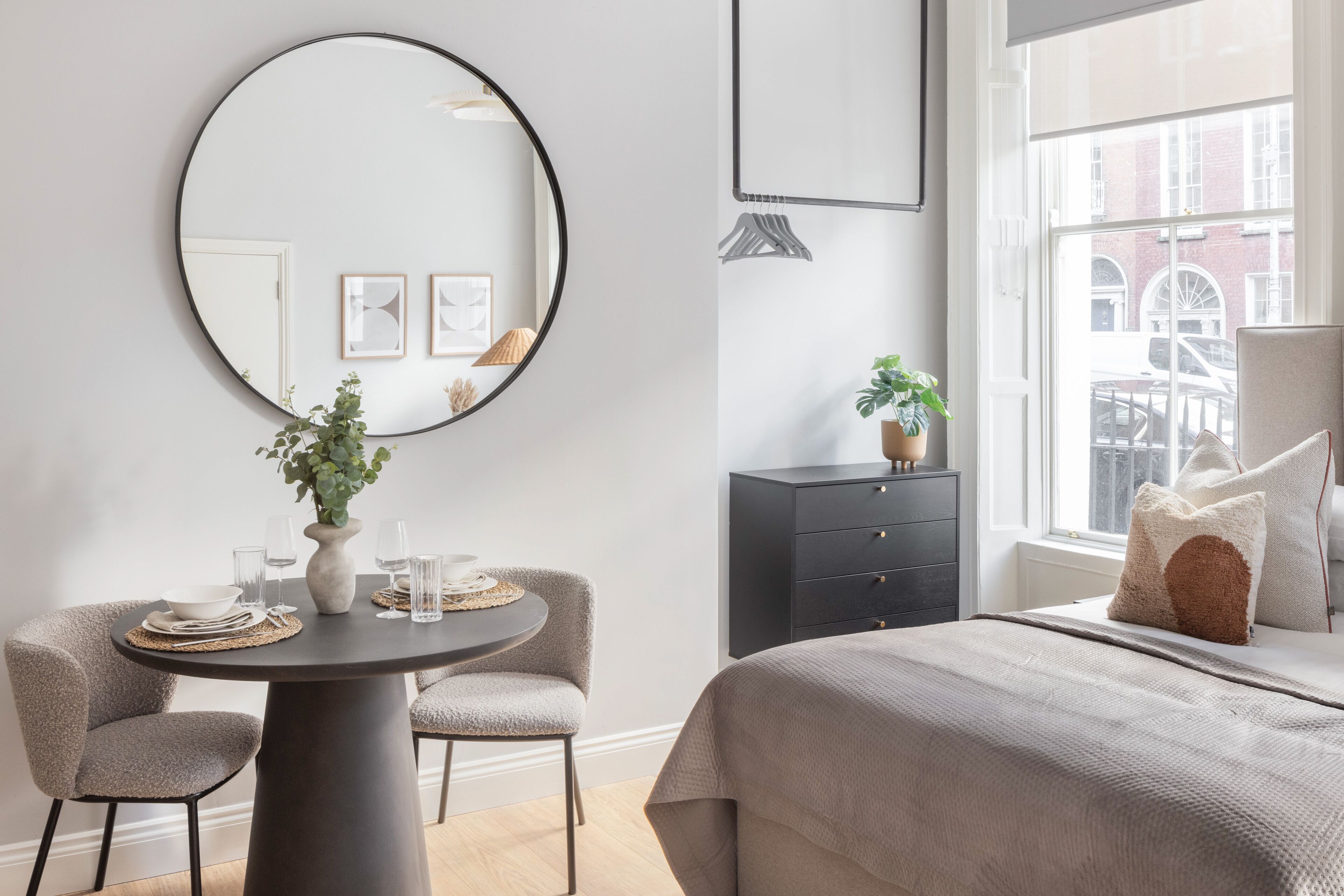A modern bedroom with a round mirror, a dining table with chairs, a bed with a textured blanket, a dresser, and a window with a view of buildings.