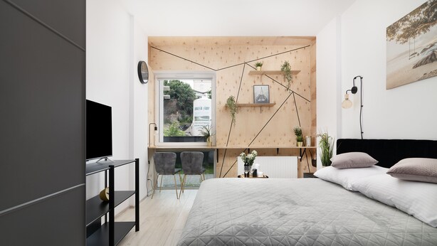 A modern bedroom with a large bed, a desk with two chairs, a window with a view of greenery, and a wall-mounted shelf with plants. - EASY RENT Apartments - Smart 005 (Lublin)