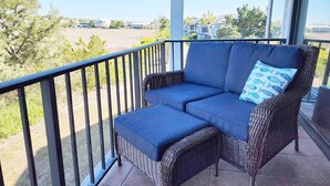A balcony with a wicker sofa and cushions, overlooking a residential area with houses and trees. - Inlet Point 14A offers Beautiful Marsh Views and Easy Creek Access (Pawleys Island)