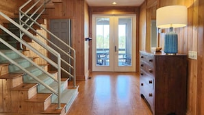Un intérieur en bois avec un escalier, une porte donnant sur l’extérieur et une commode surmontée d’une lampe.