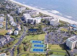 An aerial view of a coastal hotel area with multiple buildings, a beach, and tennis courts.