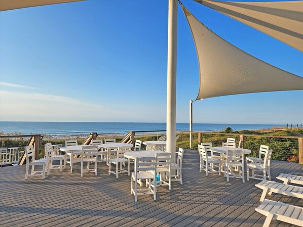 A deck with white tables and chairs, overlooking a beach and ocean.