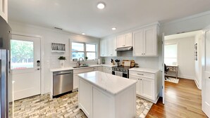 Una cocina moderna con armarios blancos, una isla central, electrodomésticos de acero inoxidable y un suelo de madera en espiga.