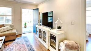 Una sala de estar con un sofá, un televisor de pantalla plana, un aparador con puertas de vidrio, un tapete estampado y una planta pequeña.